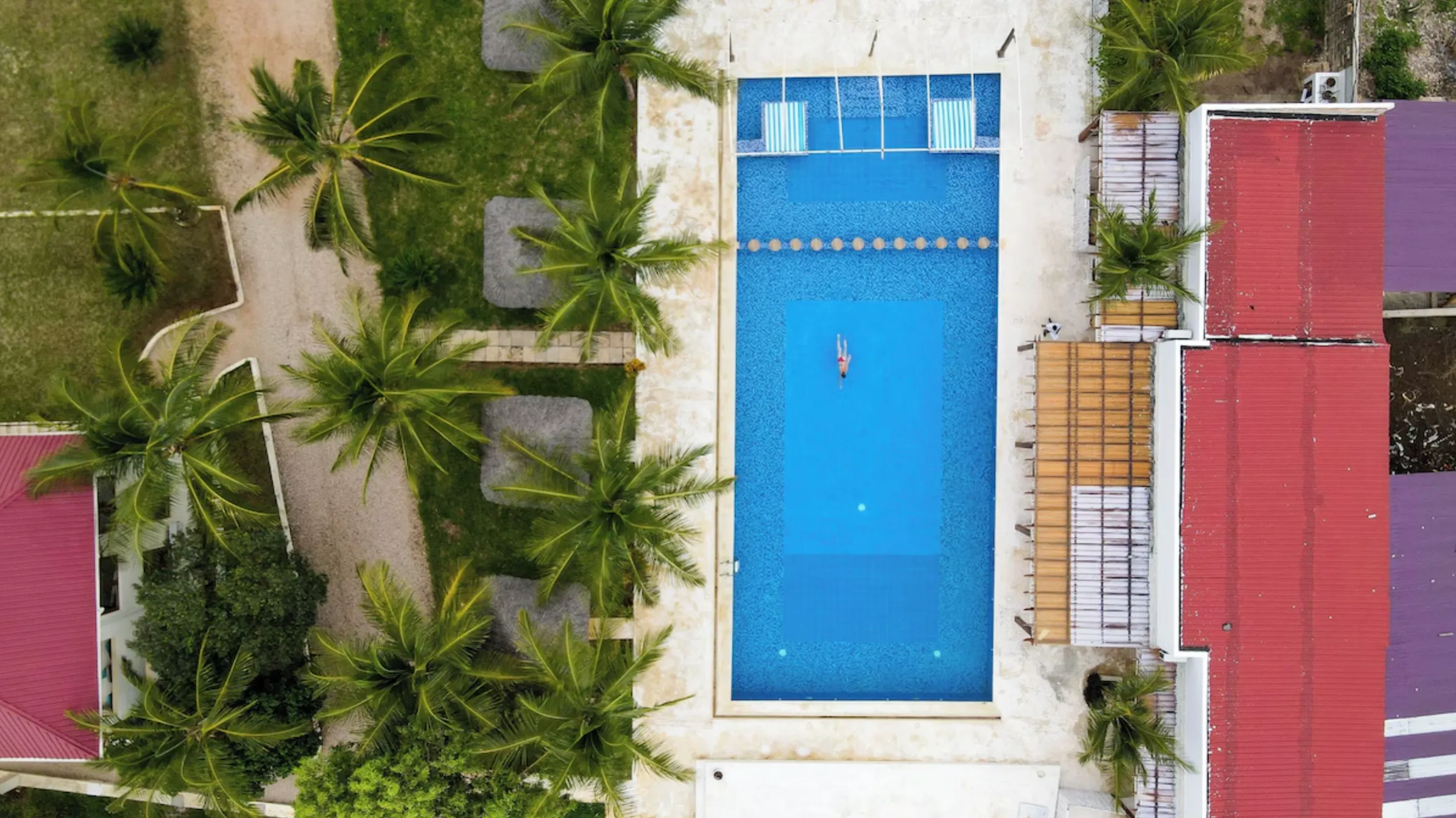 Aerial view looking straight down onto the hotel: palms, red roofs, the pool, and the empty sand beyond.